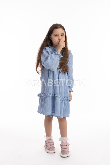 الأمر بالصمت والاستماع والتوقف عن الكلام، بورتريه لفتاة عربية خليجية اماراتية ترتدي الفستان ترفع اصبعها الى فمها, بنت سعودية تقف بإيماءات يدين تدل على طلب الهدوء و عدم الإزعاج, صورة الجسم بالكامل، خلفية بيضاء