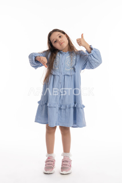 The concept of rejection and acceptance, approval and disliking, a Saudi girl standing with hand gestures indicating hesitation about something, a portrait of an Arab Emirati Gulf girl wearing a dress, raising her thumbs up and down, full-length body portrait, white background