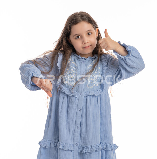 الموافقة وعدم الإعجاب، مفهوم الرفض والقبول، فتاة سعودية تقف بإيماءات يد تدل على التردد في شيء ما, بورتريه مقرب لبنت عربية اماراتية خليجية ترتدي الفستان ترفع أصابع الإبهام لأعلى وأسفل، خلفية بيضاء