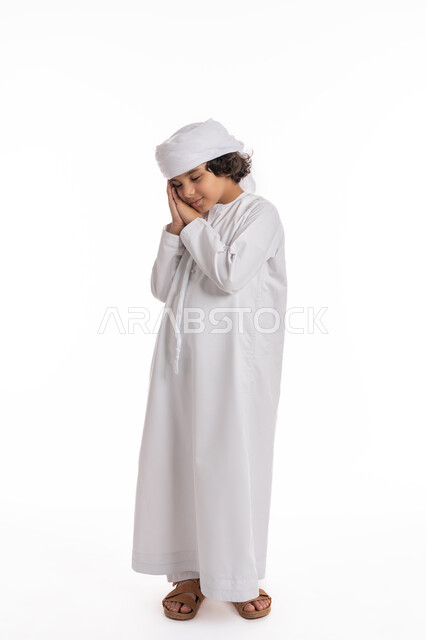 الإحساس بالكسل و الإرهاق والخمول، الشعور بالتعب و الحاجه الى النوم والراحه, بورتريه لولد عربي اماراتي خليجي يرتدي الكندورة والعمامة يضع يديه تحت رأسه, فتى سعودي يقف بإيماءات وجه ويدين تدل على النعاس, صورة الجسم بالطول الكامل، خلفية بيضاء