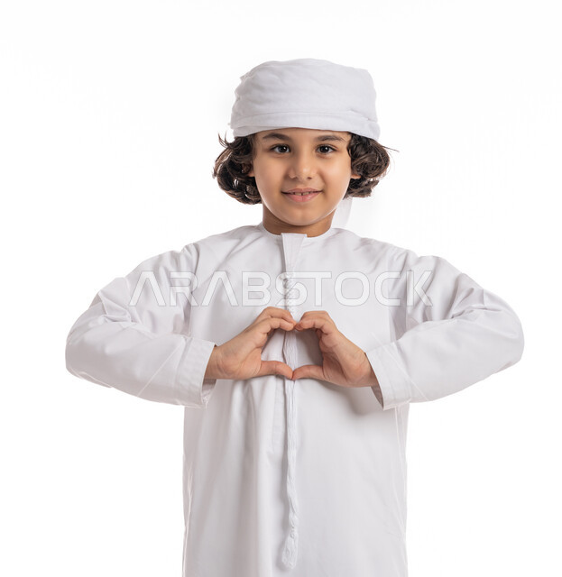 Expressing positive feelings, happiness, reassurance, and love, a close-up portrait of an Arab Emirati Gulf boy wearing a kandora and turban, raising his hands in the shape of a heart, gestures and expressions of feeling love, a Saudi boy celebrating Valentine’s Day, white background.