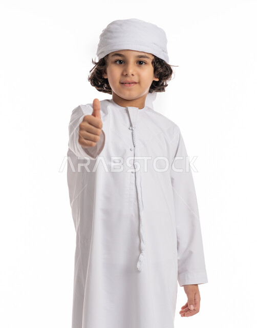 إيماءات يدين تدل على الإعجاب والرضا, بورتريه مقرب لولد عربي اماراتي خليجي يرتدي العمامة والكندورة ويشير بأصبع الابهام بعلامة الجودة, مفهوم الإمتياز والتميز, فتى سعودي يرفع أصبع الإبهام للأعلى بتعابير الموافقه والقبول، خلفية بيضاء