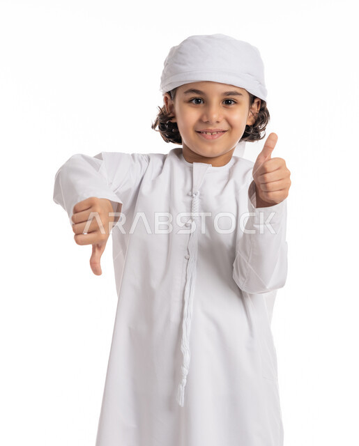 Approval and dislike, and the concept of rejection and acceptance, a Saudi boy standing with hand gestures indicating hesitation about something, a close-up portrait of an Arab-Emirati Gulf boy wearing a kandora and turban, raising his thumbs up and down, white background.