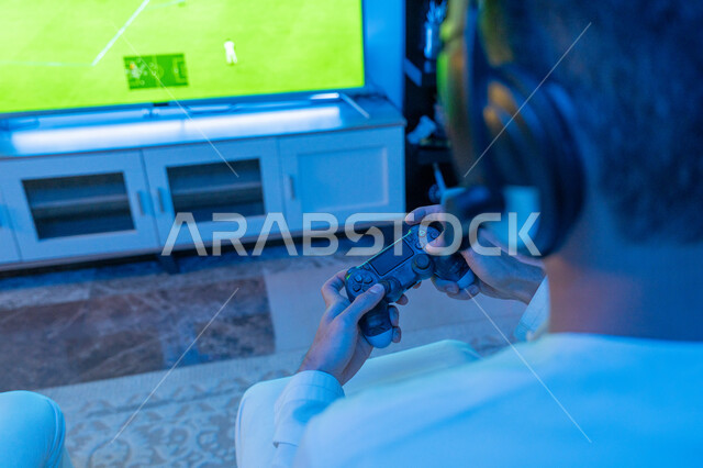The digital entertainment world, a close-up picture from the back of an Arab Gulf Emirati young man wearing the Emirati kandora and a headphone, holding a joystick in his hand, enjoying a fun and exciting entertainment experience in electronic games and modern devices, fun and entertainment with video games.
