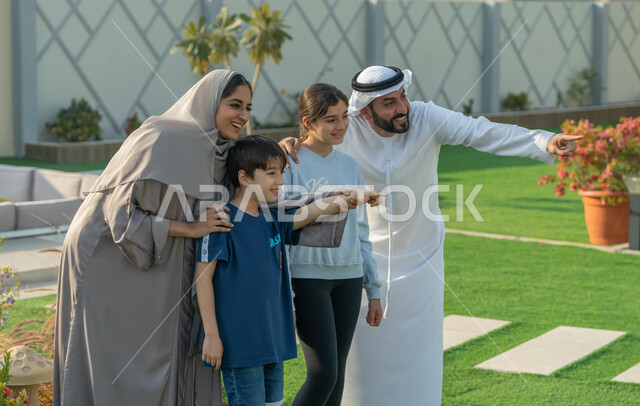 Taking a souvenir photo, documenting happy moments, the concept of family cohesion, an Arab Gulf Emirati family spending enjoyable times in one of the tourist chalets in the Emirates, hiking in the open air, an Arab Gulf Emirati father wearing a kandora and a white ghutra, pointing with his index finger to a place