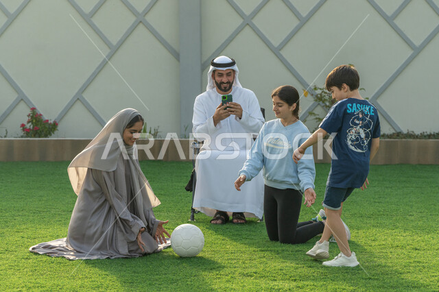 Hiking in the open air, an Arab Gulf Emirati family spending enjoyable times in one of the tourist chalets in the Emirates, an Arab Gulf Emirati father wearing a kandora and a white ghutra, taking pictures of his son playing football, spending enjoyable times with his family, a happy family atmosphere and gatherings.