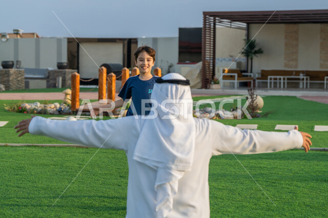 ايماءات طفل تدل على الحب و الفرح، قضاء أوقات عائلية ممتعة بالعطلة الصيفية في الحديقة, حب الآباء لأبنائهم، عناق الأبناء لأبائهم، صورة من الخلف لأب عربي خليجي اماراتي يرتدي الكندورة والغترة البيضاء يعانق إبنه بحنان, مفهوم الأسرة السعيدة