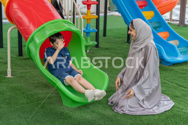 The concept of a happy family, skating on the slide, gestures of fun and happiness, using free time to have fun and play, an Arab Emirati Gulf child playing in the park, enjoying playing on a sunny day, taking a walk with his parents, spending enjoyable times in a recreational park