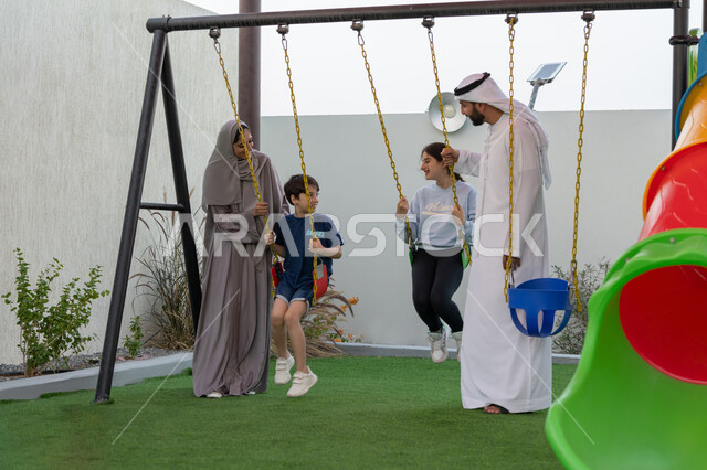 التأرجح على الأرجوحه, الإستمتاع باللعب في يوم مشمس, عائلة عربية خليجية اماراتية تقضي أوقات ممتعة في حديقة ترفيهية، قضاء الوقت مع الابناء, رجل عربي خليجي اماراتي يرتدي الكندورة والغترة البيضاء، استغلال وقت الفراغ في التسلية والمرح