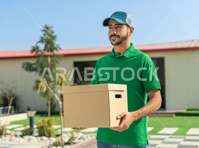 خدمة الديليفري وتوصيل الطلبات السريعة والآمنة، مندوب إماراتي خليجي عربي يرتدي الزي الكاجوال ويضع القبعة الزرقاء مبتسم يحمل طرد البريد بتعابير الرضا والسرور، الاستعداد لتسليم واستلام الطلبات