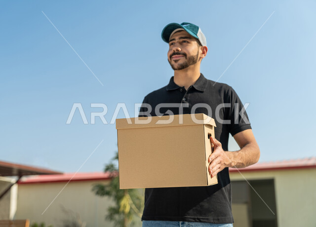 Express delivery and delivery service, an Emirati Gulf Arab worker wearing a casual outfit and wearing a blue hat, smiling, holding a mail package in his hand with an expression of satisfaction and pleasure, preparing to deliver and receive orders, high-quality shipping and delivery