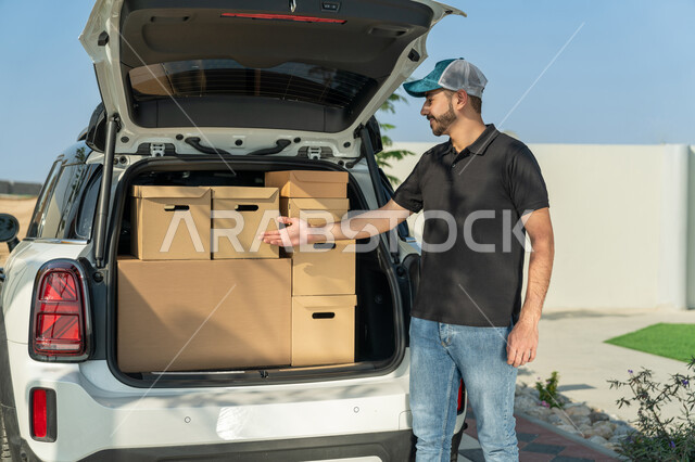 خدمة العملاء وعملية توصيل الطلبات للزبائن، مندوب إماراتي خليجي عربي يرتدي الزي الكاجوال ويضع القبعة الزرقاء يشير  إلى الصناديق الكرتونية داخل سيارة البريد، التأكد من صحة الطلبية، الشحن والتوصيل ذات الجودة العالية في الإمارات