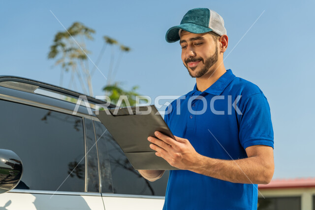 تسليم البضائع والطلبات للزبائن، عامل إماراتي خليجي عربي مبتسم يرتدي الزي الكاجوال والقبعة الزرقاء يقف بجانب سيارة البريد ويوقع على أوراق ومستندات استلام وتسليم الطرود بتعابير الرضا والثقة, خدمات التوصيل السريعة في الإمارات