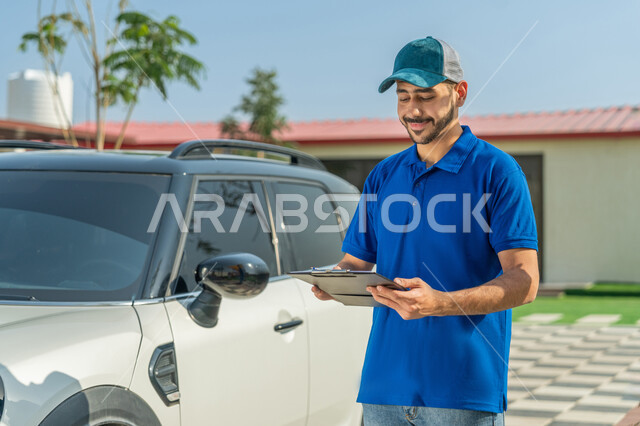 تسليم البضائع والطلبات للزبائن، عامل إماراتي خليجي عربي مبتسم يرتدي الزي الكاجوال والقبعة الزرقاء يقف بجانب سيارة البريد ويحمل أوراق ومستندات استلام وتسليم الطرود بتعابير الرضا والثقة, خدمات التوصيل السريعة في الإمارات