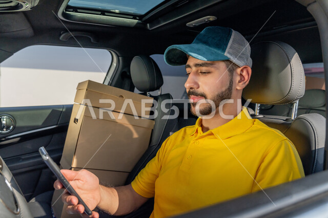 خدمات التوصيل والشحن الالكتروني الآمن, صورة مقربة لمندوب عربي خليجي إماراتي يرتدي الزي الكاجوال و القبعة الزرقاء يجلس داخل سيارة شحن البضائع ويحمل بيده الهاتف المحمول, استخدام التقنيات الحديثة في تحديد موقع العميل لتوصيل الطلبات