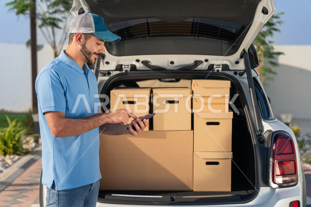 عملية الشحن الامنة, شاب عربي اماراتي خليجي يرتدي الزي كاجوال يقف بجانب سيارة شحن البضائع يحمل بيده الهاتف المحمول, استخدام التقنيات الحديثة في تحديد موقع العميل لتوصيل الطلبات, عمليات شحن وتوصيل المشتريات السريعة في الامارات, طلبات اونلاين التسوق الذكي