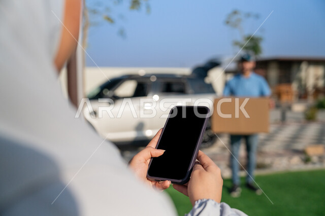 خدمة التوصيل السريع والآمن, صورة مقربة من الخلف لامرأة عربية اماراتية خليجية محجبة تحمل بيدها الهاتف المحمول للتأكد من وصول رسالة استلام الطلب, دمج التقنيات الحديثة المتطورة في الحياة العملية, عمليات الشحن والتوصيل الموثوقة , طلبات اونلاين التسوق الذكي