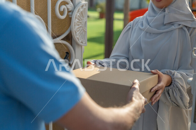 توصيل البضائع والطلبات للعميل, إمرأة عربية اماراتية خليجية محجبة ترتدي عباءة رمادية تقوم باستلام  مشترياتها منالمندوب , عمليات الشحن والتوصيل ذات الجودة العالية, خدمة الديلفري الآمنة, توصيل طلبيات الاونلاين السريع