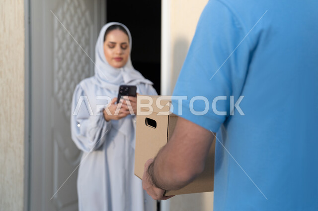 Express delivery service in the Emirates, an Emirati Gulf Arab woman wearing a gray abaya, holding a mobile phone in her hand to ensure that the order receipt message has arrived, integrating advanced modern technologies into practical life, receiving and delivering online orders, smart shopping, high-quality shipping operations
