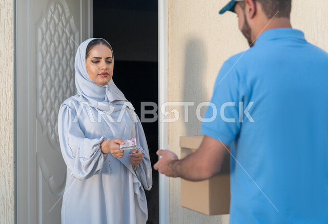 التسوق السريع الامن, امرأة عربية اماراتية خليجية محجبة ترتدي العباءة الرمادية تقوم بدفع ثمن الطلبية لمندوب التوصيل, الشحن الاونلاين السريع في الإمارات, تسليم واستلام البضائع والمشتريات, خدمة الديلفري الآمنة