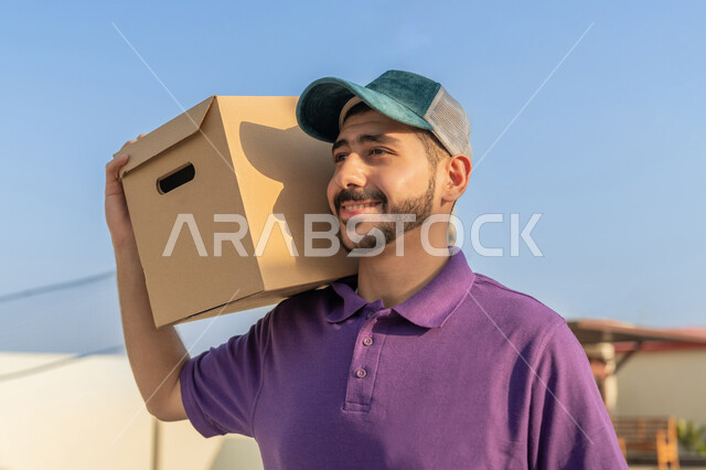خدمة الديلفري الآمنة ,  صورة مقربة لمندوب عربي اماراتي خليجي يرتدي الزي الكاجوال يحمل على كتفه طرد البريد بتعابير الرضا والسرور , عمليات الشحن والتوصيل السريعة في الامارات, الاستعداد لتسليم واستلام الطلبات
