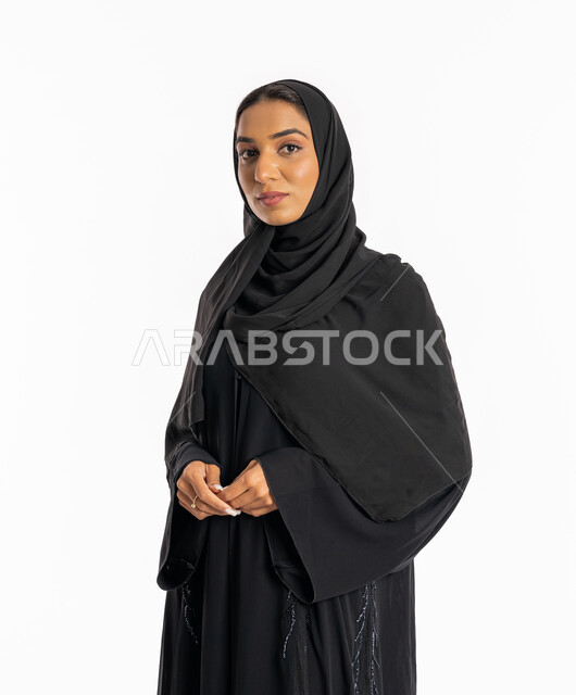 العنايه بالمظهر الخارجي، تعابير السرور والسعادة، مفهوم الأنوثة والنعومة، شابة سعودية مبتسمة تقوم بشبك يديها ببعضها، بورتريه من الجانب لامرأة عربية خليجية اماراتية محجبة ترتدي عباءة سوداء تقف باستقامة تنظر إلى الكاميرا بايماءات الثقة بالنفس، خلفية بيضاء