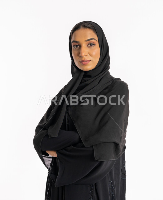 The concept of femininity and softness, a smiling young Saudi woman standing with crossed hands, attention and care for her external appearance, a portrait of a veiled Arab Gulf Emirati woman wearing a black abaya looking at the camera with gestures of self-confidence, expressions of pleasure and happiness, a white background