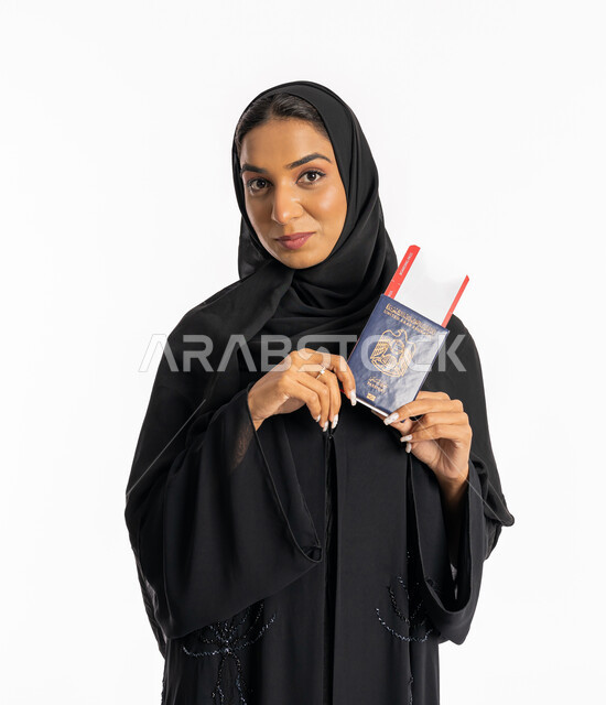 الاستعداد لقضاء رحلة صيفية ممتعة، مفهوم السياحة والسفر، القيام بالجولات الترفيهية في وقت الإجازة، بورتريه مقرب لإمرأة عربية خليجية اماراتية محجبة ترتدي عباءة سوداء تحمل بيدها الباسبور والتيكت تنظر إلى الكاميرا بإيماءات السرور، شابة سعودية أنيقة، خلفية بيضاء