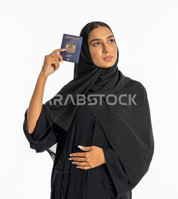 ايماءات السرور والفرح، مفهوم السياحة والسفر، القيام بالجولات الترفيهية في وقت الإجازة، بورتريه مقرب لإمرأة عربية خليجية اماراتية محجبة ترتدي عباءة سوداء ترفع الباسبور بجانب وجهها، شابة سعودية أنيقة، تعابير البهجة والسرور، خلفية بيضاء