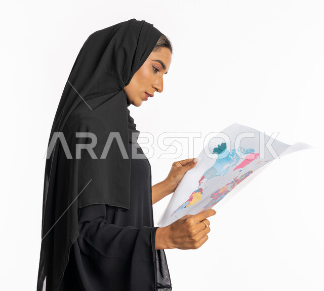 البحث والاستكشاف حول العالم، مفهوم السياحة والسفر، تحديد مسار سير الرحلة، القيام بجولات سياحية ترفيهية، بورتريه مقرب من الجانب لامرأة عربية خليجية سعودية محجبة ترتدي عباءة سوداء تحمل بيدها الخريطة، شابة سعودية انيقة، خلفية بيضاء