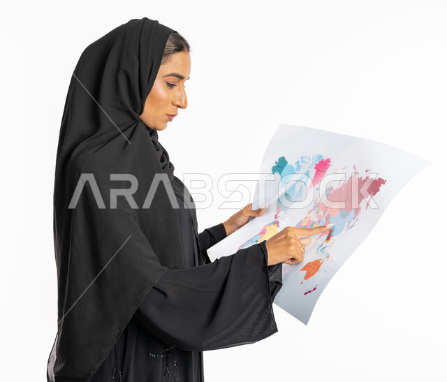 تحديد مسار سير الرحلة، البحث والاستكشاف حول العالم، مفهوم السياحة والسفر، القيام بجولات سياحية ترفيهية، بورتريه مقرب من الجانب لامرأة عربية خليجية سعودية محجبة ترتدي عباءة سوداء تحمل بيدها الخريطة، شابة سعودية انيقة، خلفية بيضاء