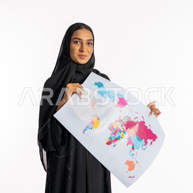 النظر إلى الكاميرا بايماءات السرور والسعادة، شابة سعودية انيقة، تحديد مسار سير الرحلة، البحث والاستكشاف حول العالم، مفهوم السياحة والسفر، القيام بجولات سياحية ترفيهية، بورتريه لامرأة عربية خليجية سعودية محجبة ترتدي عباءة سوداء تحمل بيدها الخريطة، خلفية بيضاء