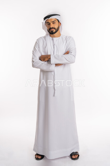 الاهتمام بالمظهر الخارجي، تعابير السرور والسعادة، مفهوم الاناقة والرجولة، النظر إلى الكاميرا بايماءات الثقة بالنفس، بورتريه لرجل عربي خليجي اماراتي يرتدي الكندورة والغرة يقف بايدي متقاطعة، صورة لكامل الجسم، خلفية بيضاء