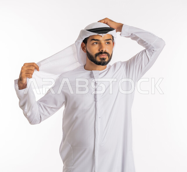 الاستعداد للخروج من المنزل، مفهوم الاناقة والرجولة، النظر إلى مكان ما بايماءات التركيز، تعابير الفرح والسعادة، الإهتمام بالمظهر الخارجي، بورتريه مقرب لرجل عربي خليجي اماراتي يرتدي الزي الاماراتي التقليدي يقوم بتعديل الغترة، خلفية بيضاء