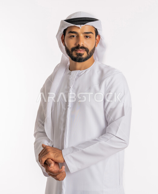 مفهوم الاناقة والرجولة، النظر إلى الكاميرا بايماءات الثقة بالنفس، تعابير السرور والسعادة، الإهتمام بالمظهر الخارجي، بورتريه لرجل عربي خليجي اماراتي يرتدي الكندورة والغترة يقف بايدي متشابكة، خلفية بيضاء