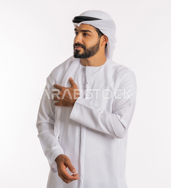 تعابير القاء التحية والسلام، النظر إلى مكان ما بايماءات التركيز، الاهتمام بالمظهر الخارجي، مفهوم الاناقة والرجولة، بورتريه لرجل عربي خليجي اماراتي يرتدي الكندورة والغرة يقف بايدي متقاطعة، خلفية بيضاء