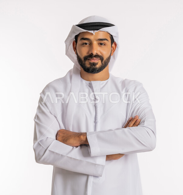 تعابير السرور والسعادة، الاهتمام بالمظهر الخارجي، مفهوم الاناقة والرجولة، النظر إلى الكاميرا بايماءات الثقة بالنفس، بورتريه مقرب لرجل عربي خليجي اماراتي يرتدي الكندورة والغرة يقف بايدي متقاطعة، خلفية بيضاء