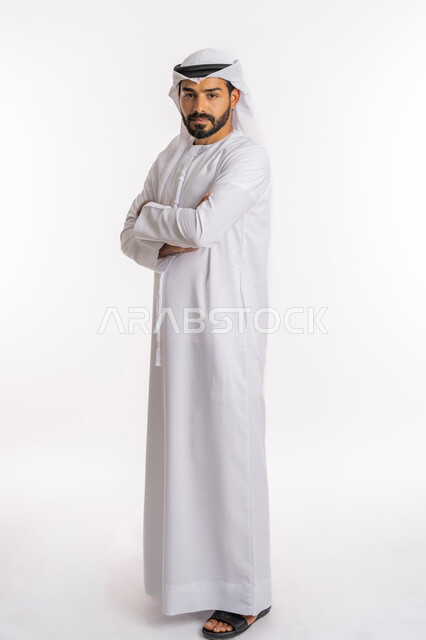 النظر إلى الكاميرا بايماءات الثقة بالنفس، الاهتمام بالمظهر الخارجي، تعابير السرور والسعادة، مفهوم الاناقة والرجولة، بورتريه من الجانب لرجل عربي خليجي اماراتي يرتدي الكندورة والغرة يقف بايدي متقاطعة، صورة لكامل الجسم، خلفية بيضاء