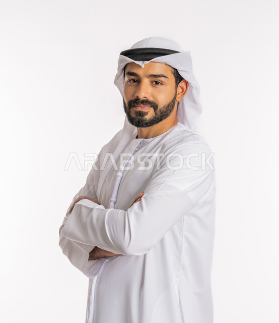 تعابير السرور والسعادة، الاهتمام بالمظهر الخارجي، مفهوم الاناقة والرجولة، النظر إلى الكاميرا بايماءات الثقة بالنفس، بورتريه مقرب من الجانب لرجل عربي خليجي اماراتي يرتدي الكندورة والغرة يقف بايدي متقاطعة، خلفية بيضاء