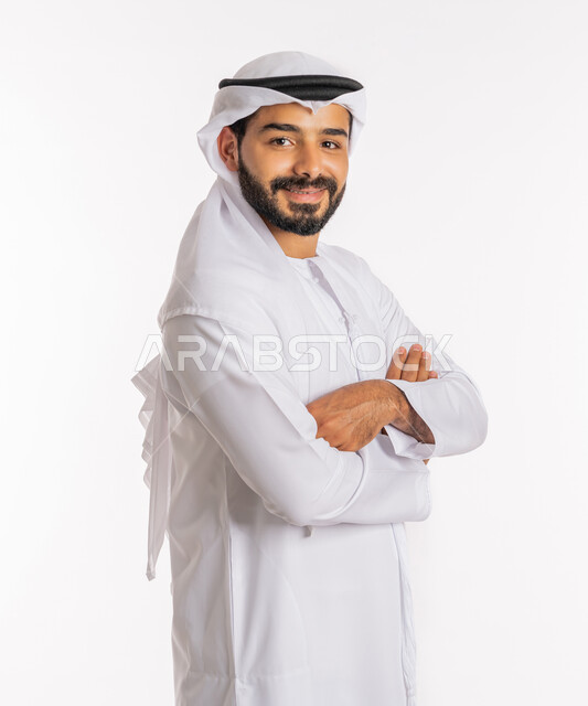 الاهتمام بالمظهر الخارجي، تعابير السرور والسعادة، مفهوم الاناقة والرجولة، النظر إلى الكاميرا بايماءات الثقة بالنفس، بورتريه مقرب لرجل عربي خليجي اماراتي يرتدي الكندورة والغرة يقف بايدي متقاطعة، خلفية بيضاء
