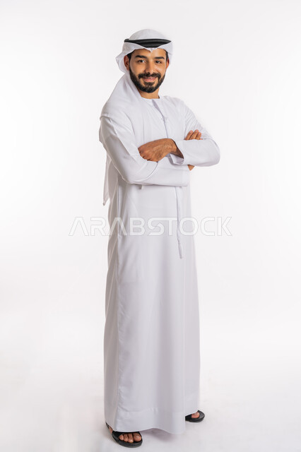 النظر إلى الكاميرا بايماءات الثقة بالنفس، الاهتمام بالمظهر الخارجي، تعابير السرور والسعادة، مفهوم الاناقة والرجولة، بورتريه من الجانب لرجل عربي خليجي اماراتي يرتدي الكندورة والغرة يقف بايدي متقاطعة، صورة لكامل الجسم، خلفية بيضاء