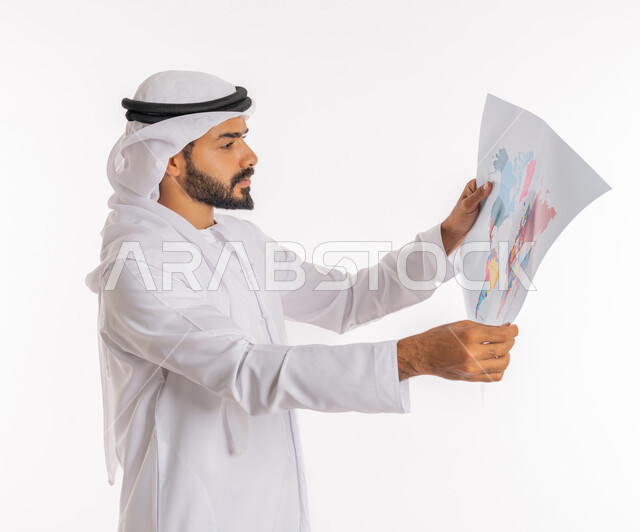 تحديد مسار سير الرحلة، البحث والاستكشاف حول العالم، مفهوم السياحة والسفر، القيام بجولات سياحية ترفيهية، بورتريه مقرب من الجانب لرجل عربي خليجي اماراتي يرتدي الكندورة والغترة يحمل بيده الخريطة، خلفية بيضاء
