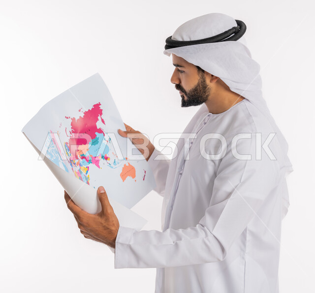تحديد مسار سير الرحلة، البحث والاستكشاف حول العالم، مفهوم السياحة والسفر، القيام بجولات سياحية ترفيهية، بورتريه مقرب من الجانب لرجل عربي خليجي اماراتي يرتدي الكندورة والغترة يحمل بيده الخريطة، خلفية بيضاء