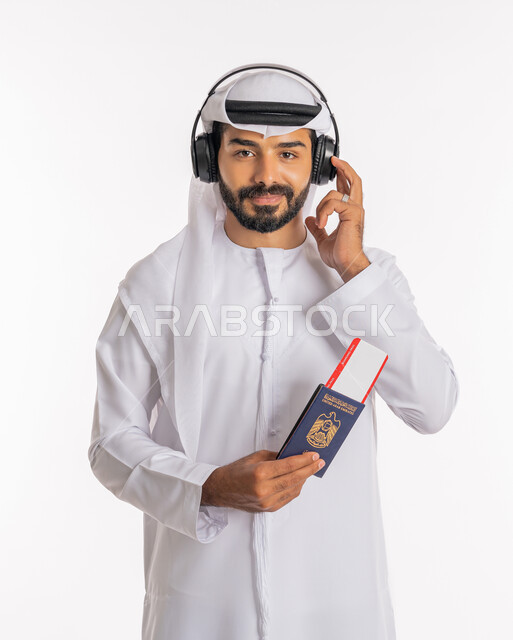 إيماءات الفرح والسعادة، مفهوم السياحة والسفر، القيام بالجولات الترفيهية في وقت الإجازة، تأشيرة سفر دولية، بورتريه لرجل عربي خليجي اماراتي يرتدي الكندورة والغترة والسماعات يحمل بيده الباسبور وتيكت الطيارة، خلفية بيضاء