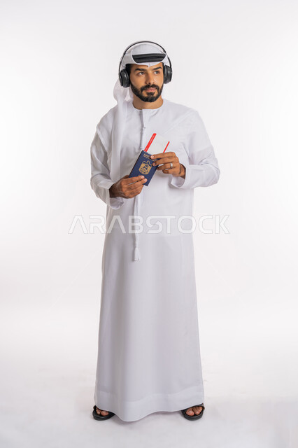 القيام بالجولات الترفيهية في وقت الإجازة، مفهوم السياحة والسفر، إيماءات الفرح والسعادة، تأشيرة سفر دولية، بورتريه لرجل عربي خليجي اماراتي يرتدي الكندورة والغترة والسماعات يحمل بيده الباسبور وتيكت الطيارة، صورة لكامل الجسم، خلفية بيضاء