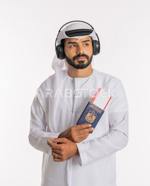 تأشيرة سفر دولية، إيماءات الفرح والسعادة، مفهوم السياحة والسفر، القيام بالجولات الترفيهية في وقت الإجازة، بورتريه مقرب لرجل عربي خليجي اماراتي يرتدي الكندورة والغترة والسماعات يحمل بيده الباسبور وتيكت الطيارة، خلفية بيضاء