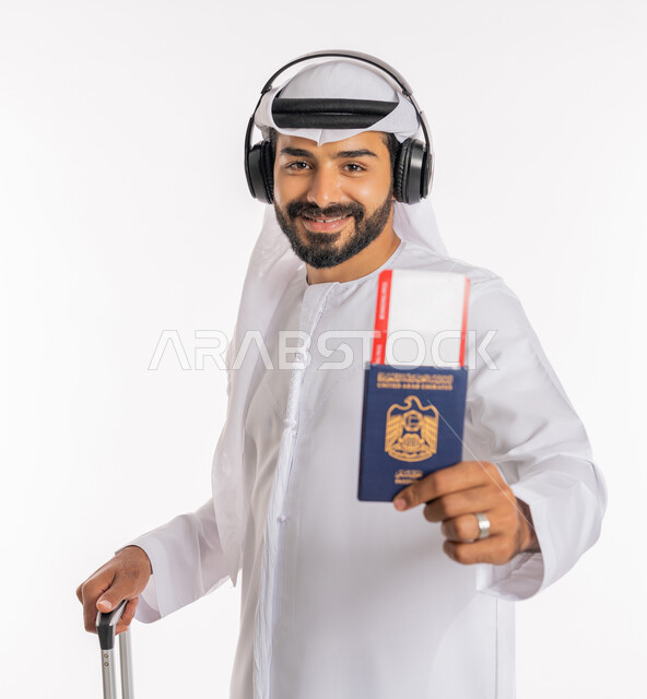 تأشيرة سفر دولية، إيماءات الفرح والسعادة، مفهوم السياحة والسفر، القيام بالجولات الترفيهية في وقت الإجازة، بورتريه مقرب لرجل عربي خليجي اماراتي يرتدي الكندورة والغترة والسماعات يحمل بيده الباسبور وتيكت الطيارة يمسك بحقيبة السفر، خلفية بيضاء
