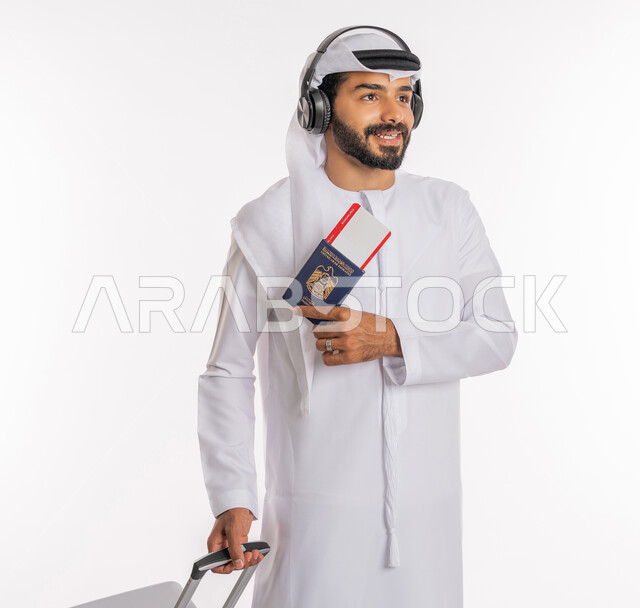 مفهوم السياحة في إجازة الصيف، الاستعداد لقضاء رحلة صيفية ممتعة، القيام بالجولات الترفيهية، بورتريه لرجل عربي خليجي اماراتي يرتدي الكندورة والغترة وسماعة الرأس يحمل بيده جواز السفر وتيكت الطيارة يمسك بحقيبة الامتعة، خلفية بيضاء
