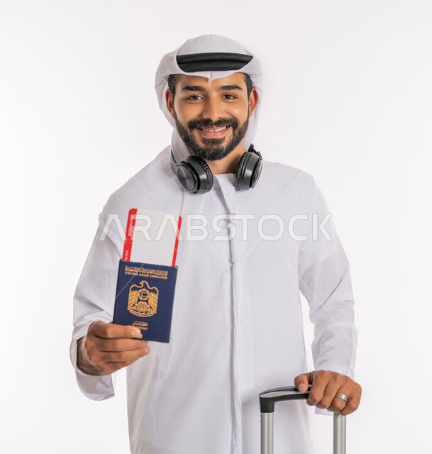 إيماءات السرور والسعادة، القيام بالجولات الترفيهية، مفهوم السياحة في إجازة الصيف، الاستعداد لقضاء رحلة صيفية ممتعة، بورتريه مقرب لرجل عربي خليجي اماراتي يرتدي الكندورة والغترة وسماعة الرأس يحمل بيده جواز السفر وتيكت الطيارة يمسك بحقيبة الامتعة، خلفية بيضاء