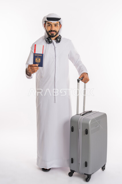 الاستعداد لقضاء رحلة صيفية ممتعة، القيام بالجولات الترفيهية، مفهوم السياحة في إجازة الصيف، بورتريه لرجل عربي خليجي اماراتي يرتدي الكندورة والغترة وسماعة الرأس يحمل بيده جواز السفر وتيكت الطيارة يمسك بحقيبة الامتعة، صورة لكامل الجسم، خلفية بيضاء